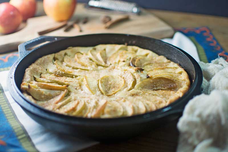 A German Apple Pancake sits baked in a black cast-iron skillet, topped with sliced apples. In the background, apples, cinnamon sticks, a grater, and a wooden cutting board add to the cozy kitchen scene.