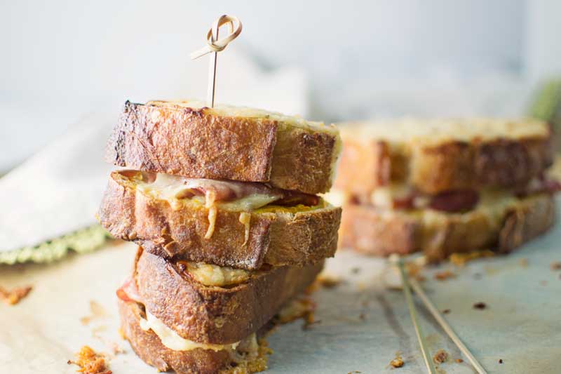 Two stacked grilled cheese sandwiches, reminiscent of a classic Croque Monsieur, with melted cheese and crispy bread, held together by a wooden skewer. Another sandwich rests on parchment paper in the background.