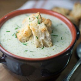 A bowl of Creamy Broccoli Potato Soup garnished with chopped chives and three pieces of toasted bread, served in a dark ceramic bowl with a spoon beside it on a wooden surface.