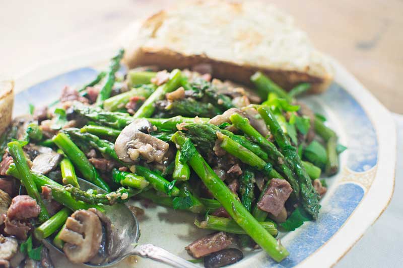 A plate of sautéed asparagus mushrooms with prosciutto is served alongside a slice of buttered bread. A spoon rests on the plate next to the vegetables.