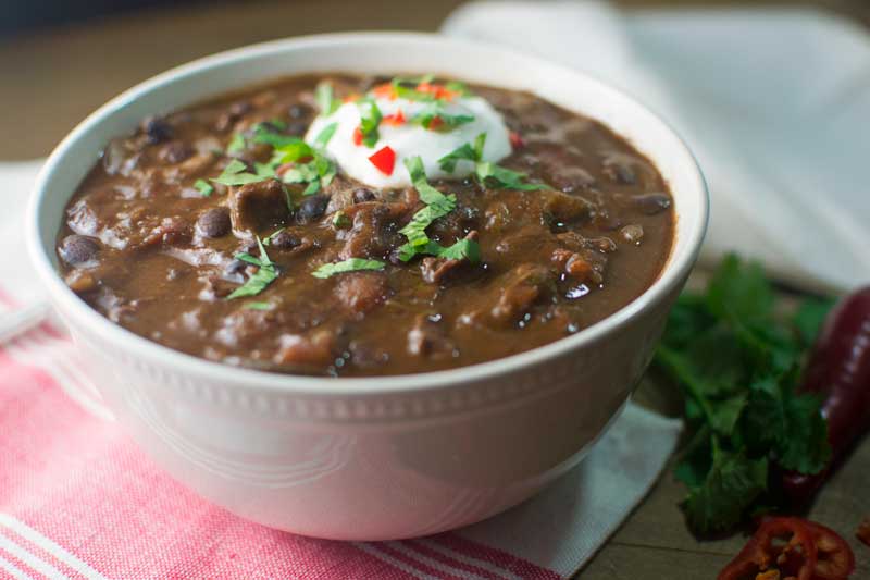 A bowl of Slow Cooker Chili topped with a dollop of sour cream, chopped cilantro, and diced red peppers rests on a red and white striped cloth, with fresh herbs in the background.