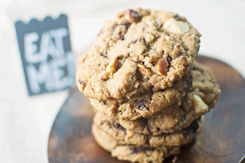 A close-up of a stack of spiced chocolate chunk cookies with visible nuts on a wooden plate. In the blurred background, a small sign reads EAT ME.