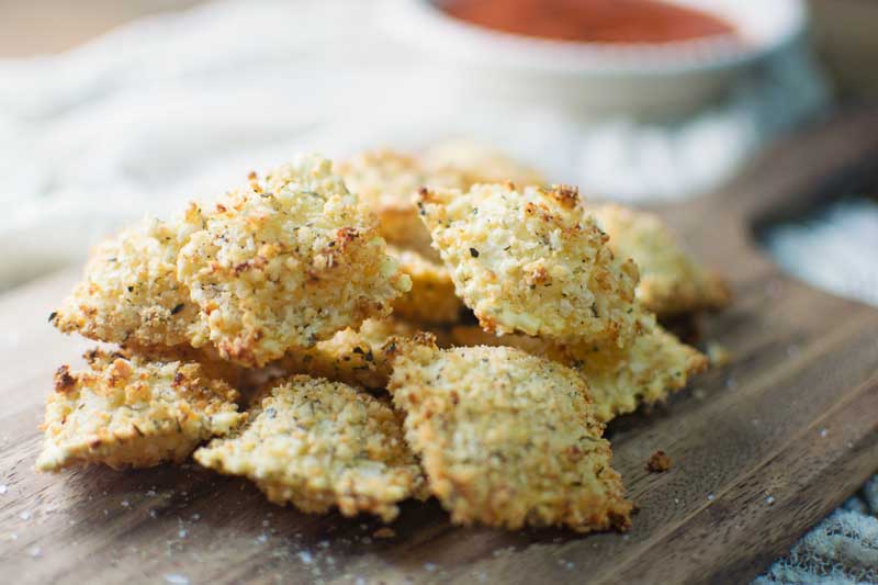 A pile of golden, crispy toasted ravioli sits on a wooden board, dusted with seasoning. A bowl of red dipping sauce is blurred in the background.