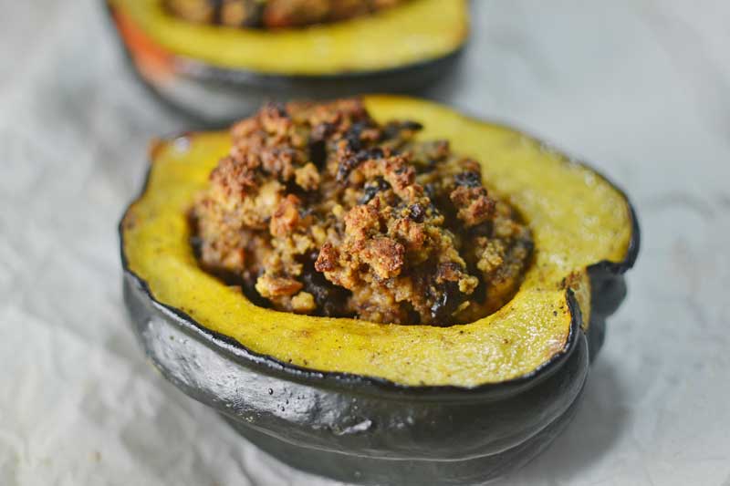 A Roasted Stuffed Acorn Squash half filled with browned, crumbly stuffing sits on a light surface, while another stuffed squash half is blurred in the background.
