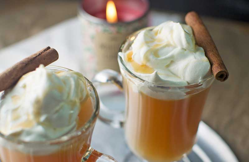 Two glass mugs filled with warm Mulled Apple Cider topped with whipped cream and a drizzle of syrup, each garnished with a cinnamon stick. A lit candle in the background adds a cozy ambiance.