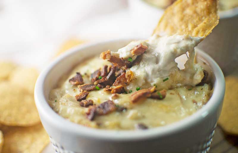 A close-up of Caramelized Onion Bacon Dip topped with chopped bacon and chives in a white bowl, with a tortilla chip scooping some dip. Other chips are blurred in the background.