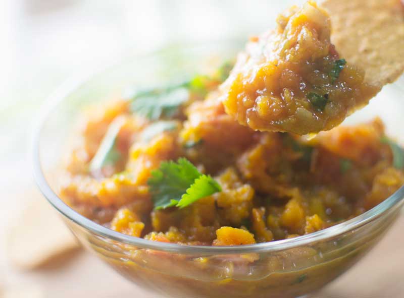 A close-up of a clear glass bowl filled with chunky Roasted Butternut Salsa, garnished with fresh cilantro, with a chip scooping some salsa from the bowl.