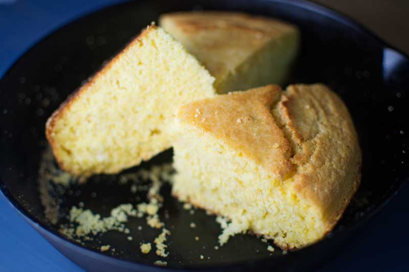 A close-up of two thick, golden slices of skillet cornbread in a black skillet, with crumbs scattered around. The skillet cornbread looks moist and crumbly with a slightly browned top.