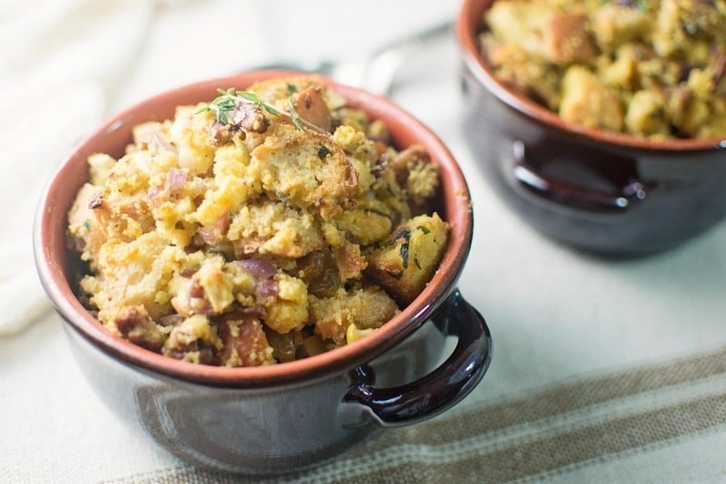 Two small dark bowls filled with golden brown cornbread dressing made of bread cubes, herbs, and bits of vegetables, garnished with fresh thyme, sit on a light-colored surface.