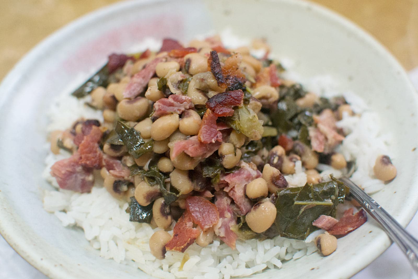 A bowl of white rice topped with savory Black Eyed Peas, collard greens, and pieces of ham, served with a fork on the side.