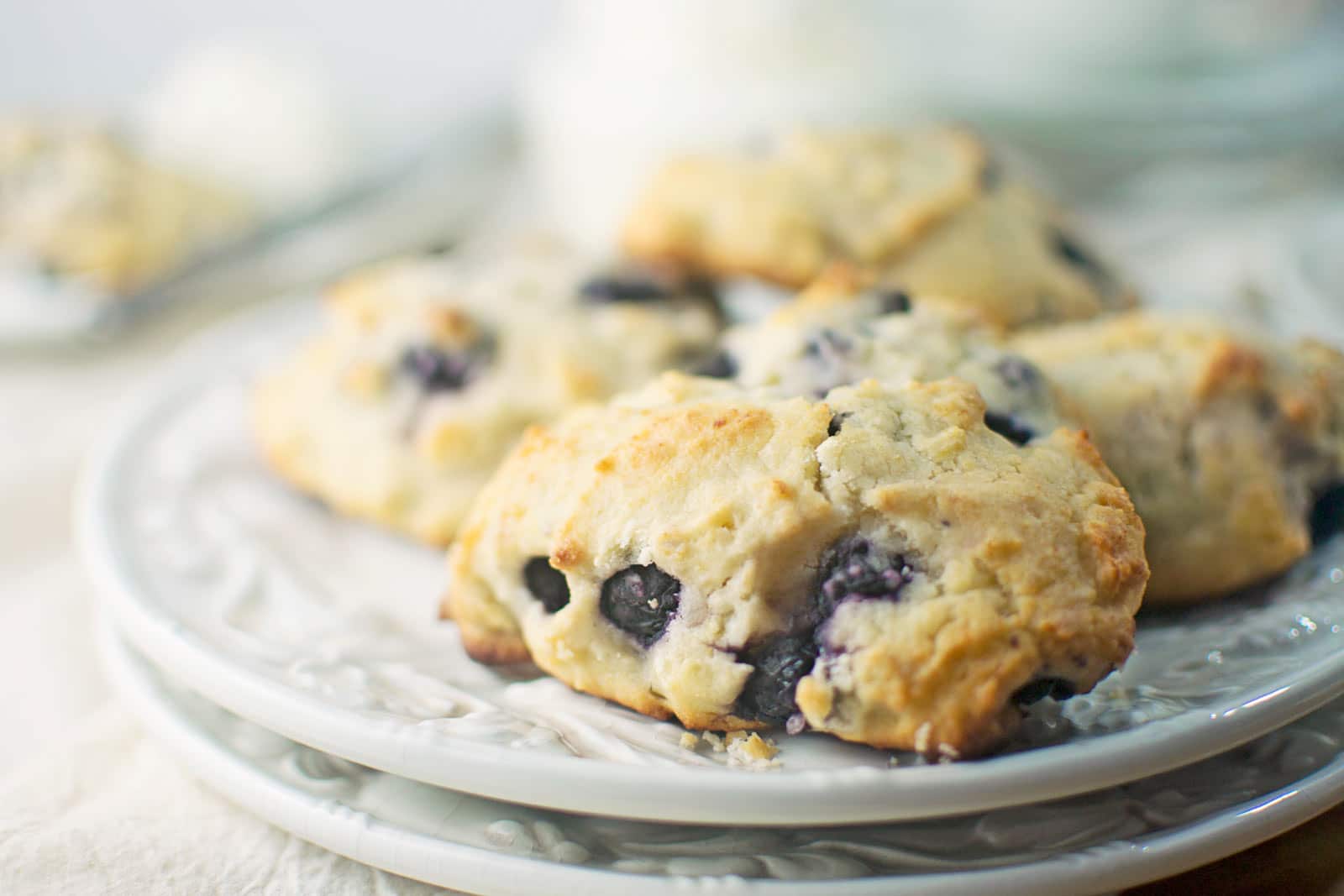 A close-up of freshly baked Blueberry Lemon Ricotta Scones on a stack of white plates, with golden-brown tops and visible blueberries. The softly blurred background highlights the scones in the foreground.