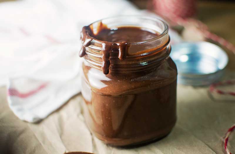 An open glass jar filled with thick, creamy Chocolate Chestnut Cream, with some sauce dripping down the rim. The jar sits on a piece of brown paper, while a lid and a white cloth appear softly in the background.