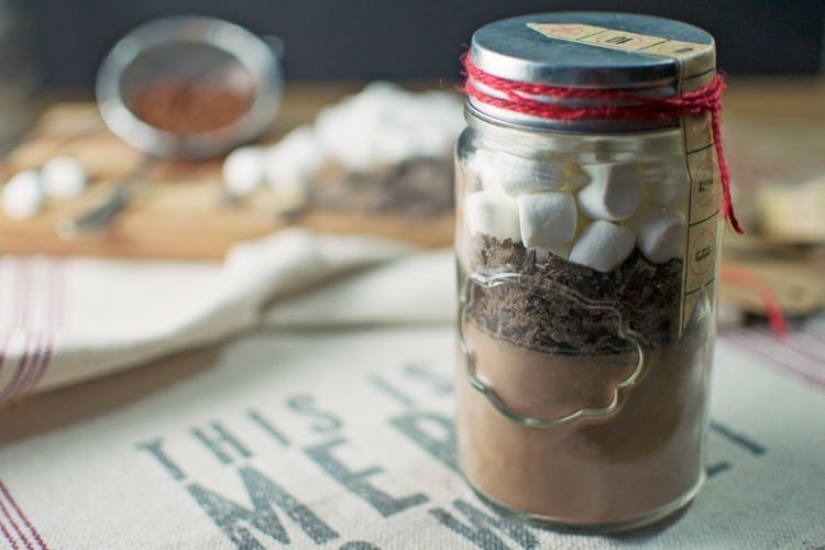 A mason jar filled with layered Hot Cocoa Mix, chocolate shavings, and mini marshmallows, topped with a metal lid and red twine, sits on a cloth with partially visible text. In the blurred background are scattered marshmallows and cocoa.