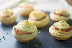 Six savory Rustic Potato Tarts topped with mashed potatoes, herbs, and a slice of ham are arranged on a dark slate surface, garnished with scattered sprigs of fresh thyme.