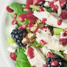 A plate of salad with mixed greens, blackberries, walnuts, shredded chicken, crumbled cheese, and a drizzle of Blackberry Balsamic Vinaigrette. A fork rests on the plate’s edge.