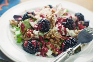 A fresh salad with blackberries, walnuts, feta cheese, greens, and a drizzle of Blackberry Balsamic Vinaigrette is served on a white plate with a fork.