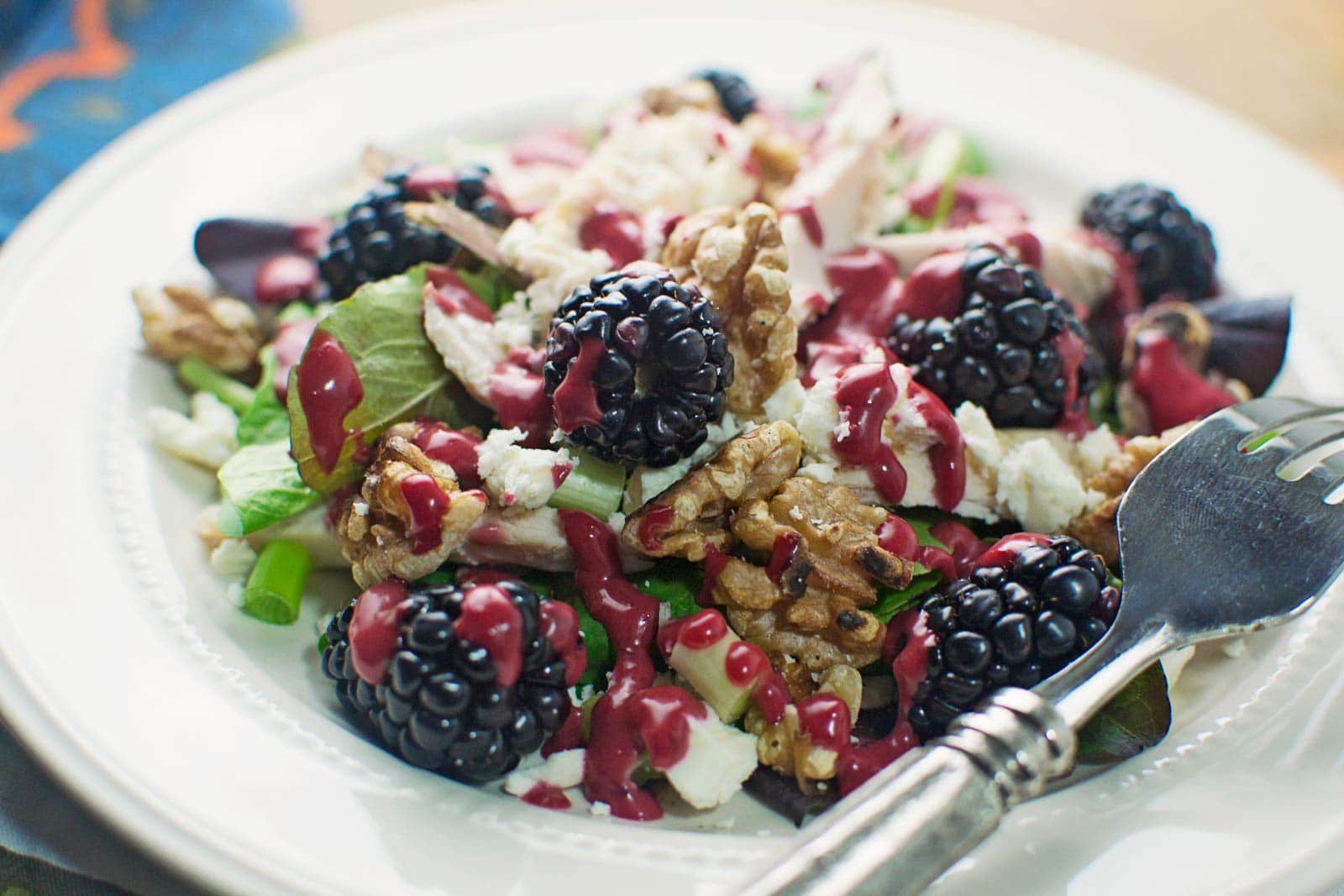 A fresh salad with blackberries, walnuts, feta cheese, greens, and a drizzle of Blackberry Balsamic Vinaigrette is served on a white plate with a fork.