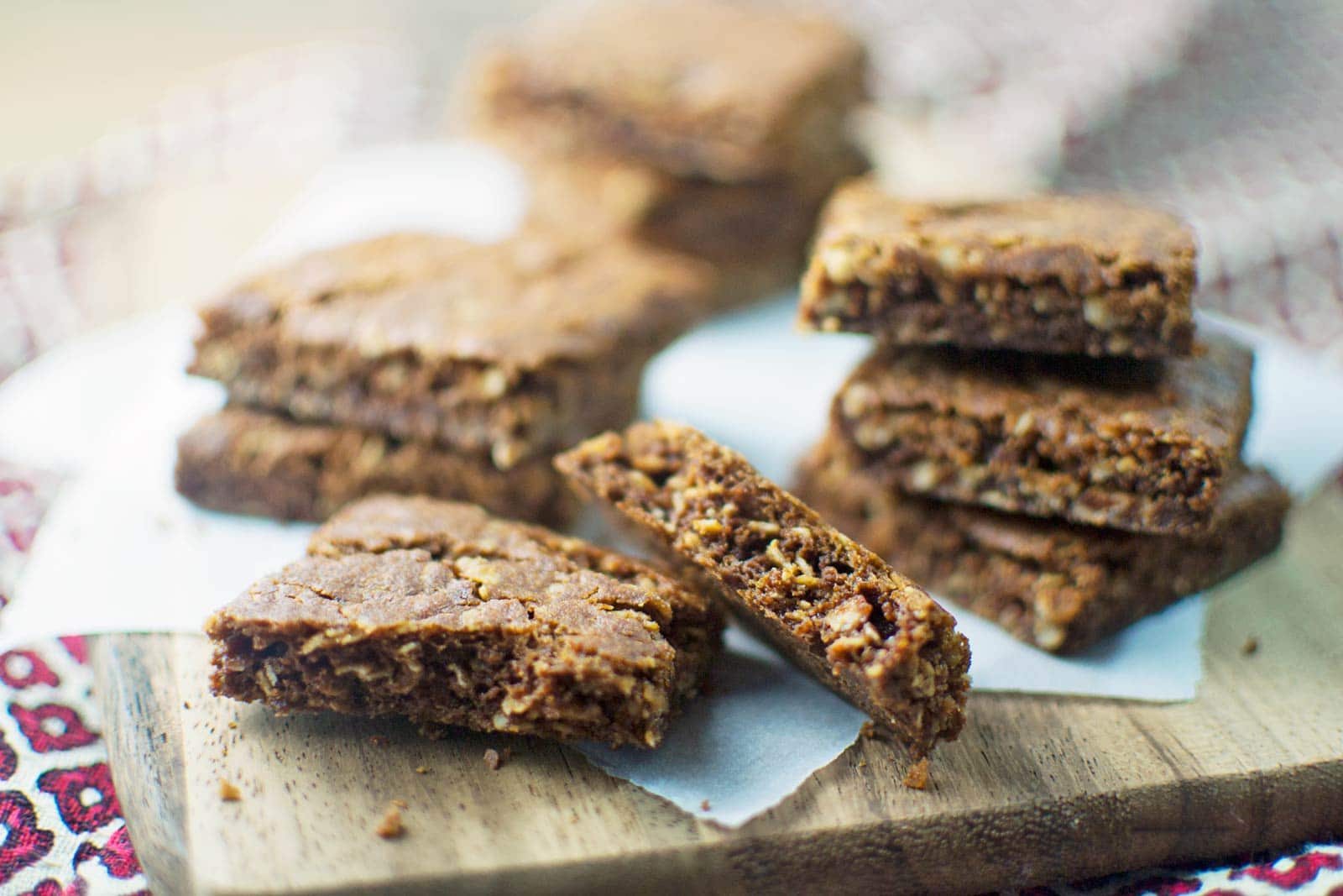 Several chewy, golden-brown Ginger Snap cookie bars are stacked and scattered on a wooden board lined with parchment paper. The bars have a crumbly texture and appear moist inside, with some pieces broken to reveal the gingery interior.