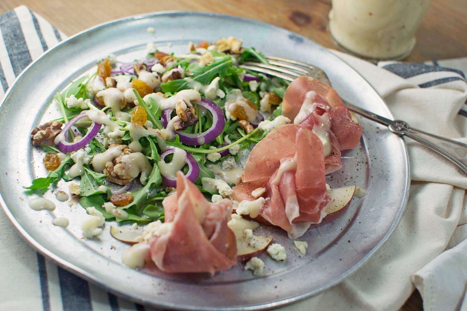 A metal plate with arugula salad topped with red onions, walnuts, golden raisins, crumbled cheese, and a creamy Honey Pear Vinaigrette, served with slices of prosciutto and a fork on a striped napkin.