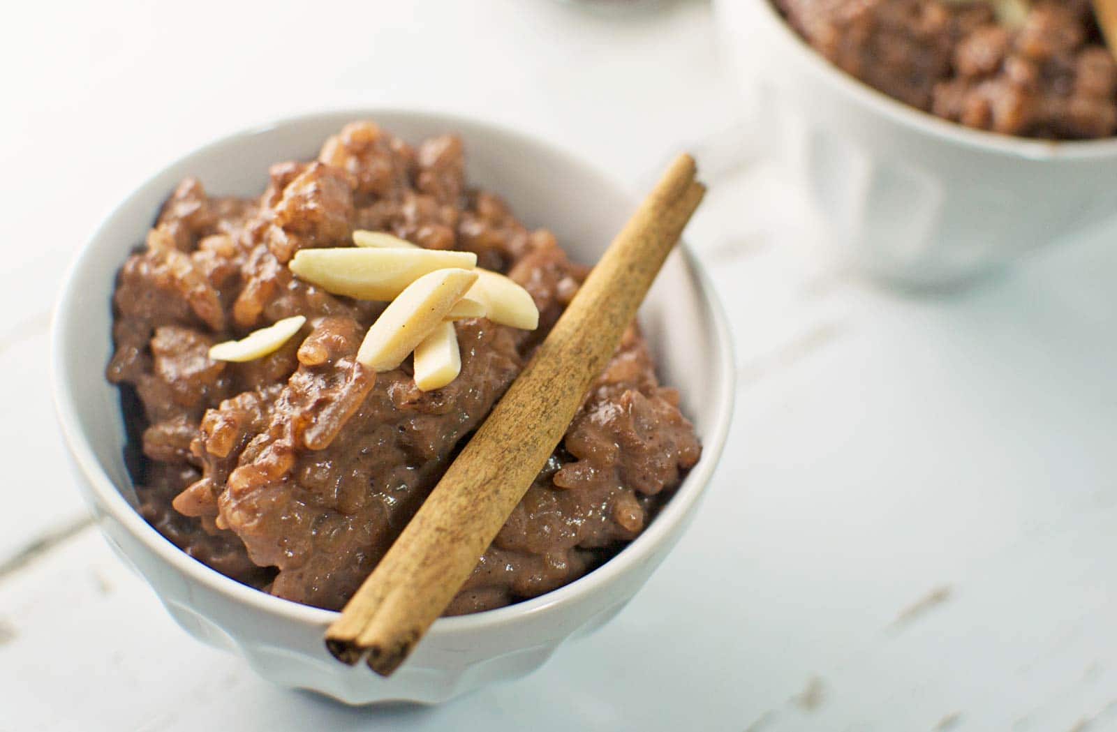 A white bowl filled with creamy chocolate rice pudding, topped with slivered almonds and a cinnamon stick, sits on a white surface. Another delicious bowl of rice pudding is blurred in the background.