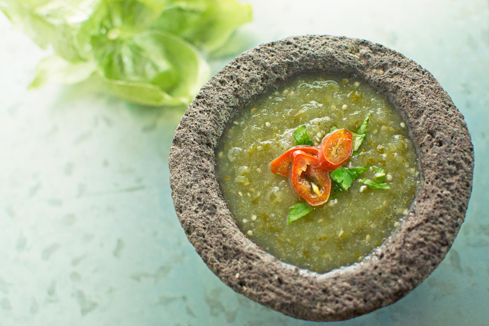 A stone bowl filled with vibrant salsa verde, garnished with sliced red chili and cilantro, with fresh tomatillos in the background on a light surface.