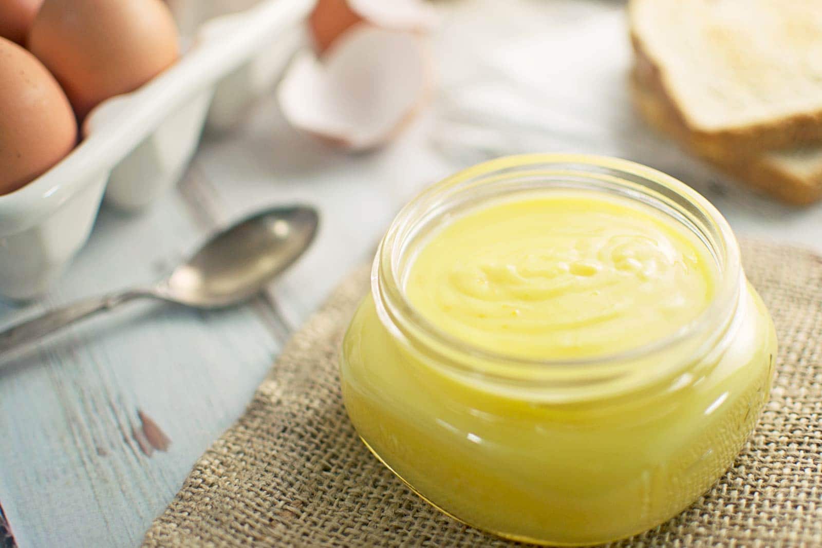 A small glass jar filled with creamy yellow lemon curd sits on a piece of burlap, with eggs, a spoon, and slices of bread in the blurred background.