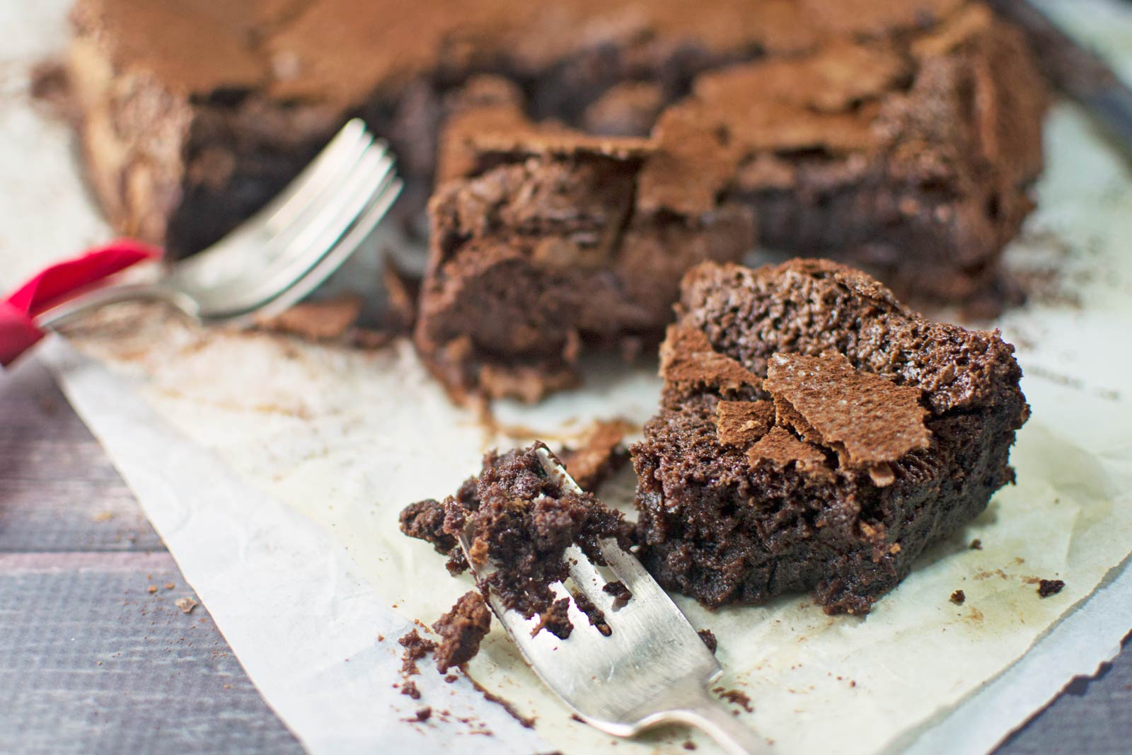 A rich, fudgy Chocolate Cake with a cracked top sits on parchment paper. One slice is partially cut with a fork, and another fork rests nearby. Chocolate crumbs are scattered on the surface.
