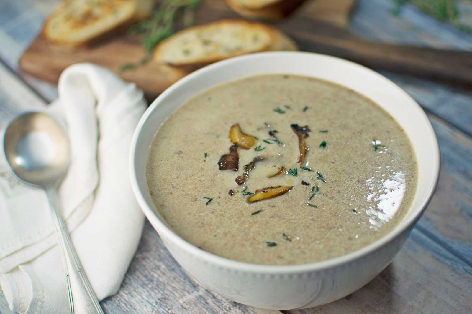 A bowl of creamy Mushroom Soup garnished with mushroom slices and herbs sits on a table next to a spoon, a white napkin, and slices of toasted bread on a wooden board in the background.