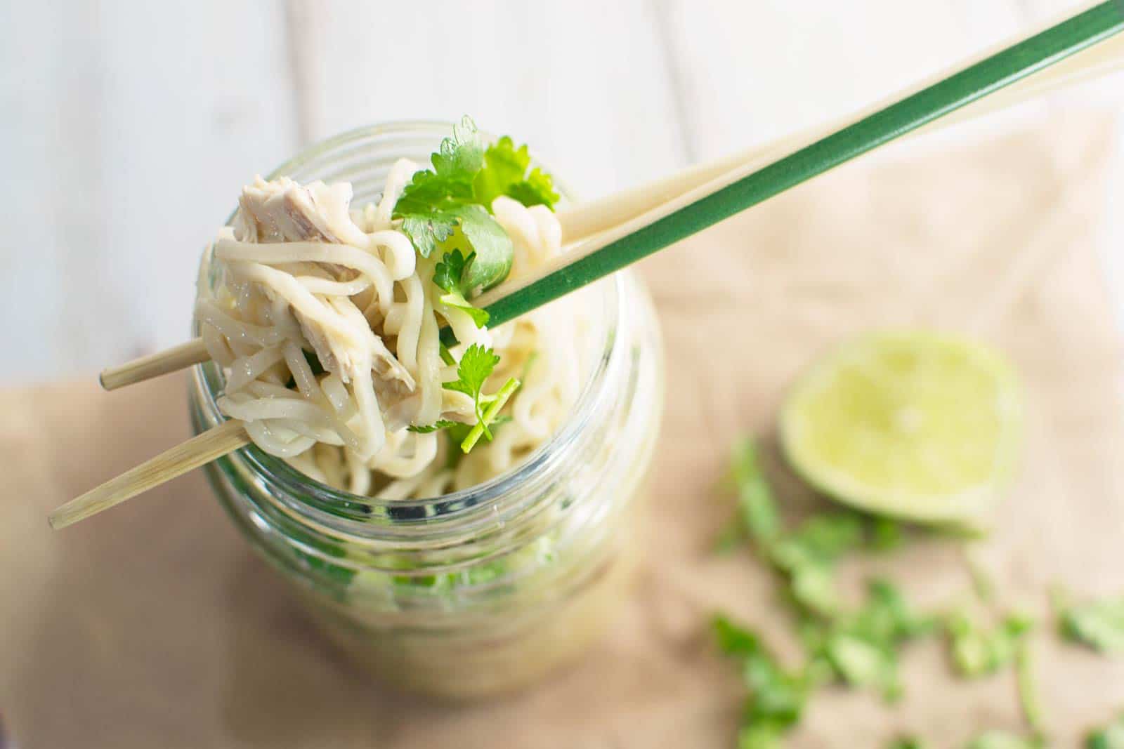 A close-up of chopsticks lifting noodles from a glass jar filled with Pot Noodles soup, garnished with cilantro. A sliced lime and scattered cilantro leaves are visible in the blurred background.