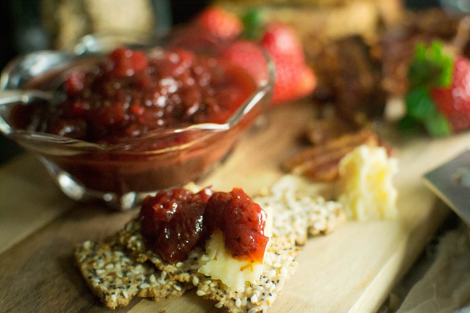 A seeded cracker topped with cheese and strawberry jam sits on a wooden board, creating a delightful cheeseboard scene beside a bowl of jam and fresh strawberries in the background.