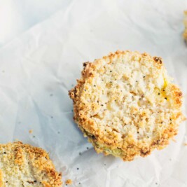 Three crispy, round, breaded and baked slices of food, possibly green tomatoes or eggplant, are placed on a sheet of white parchment paper, with a light and airy texture visible on their golden-brown edges.
