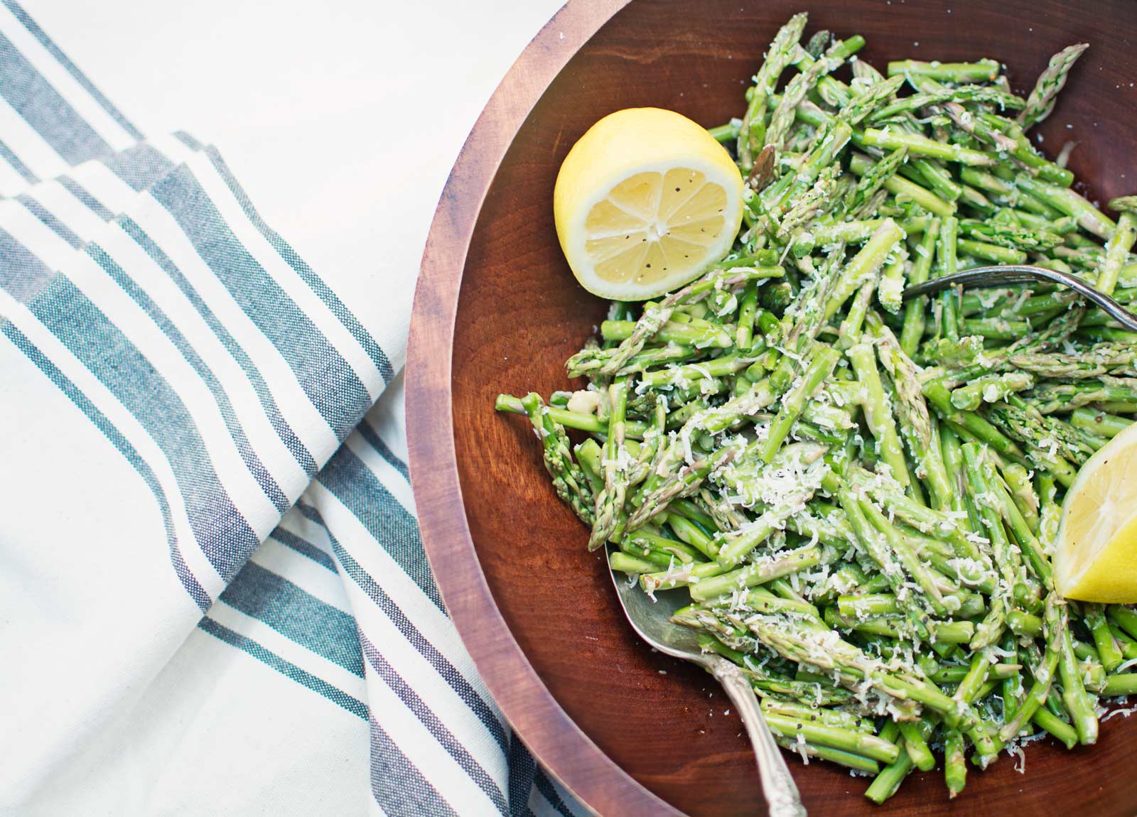 A wooden bowl filled with sautéed asparagus, grated cheese, and lemon wedges sits on a striped cloth. Two metal serving spoons rest in the bowl, ready to serve this vibrant asparagus dish.