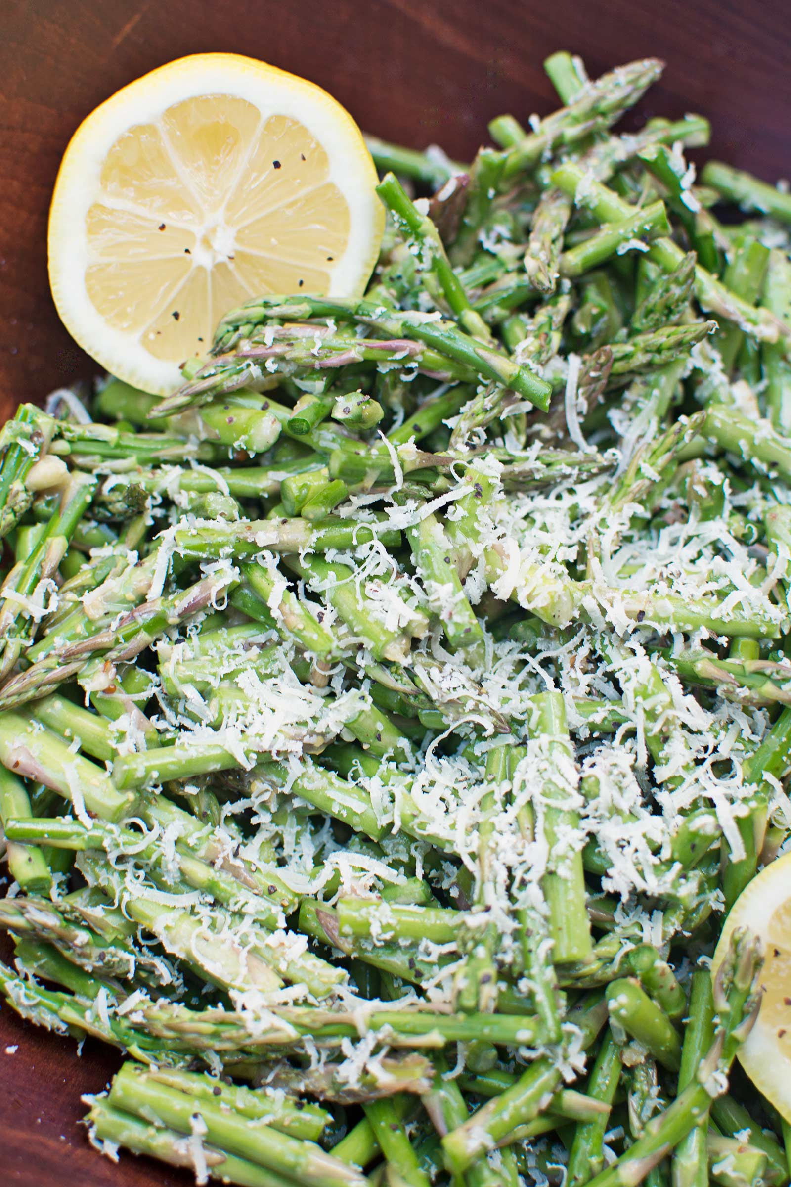 Chopped asparagus topped with grated cheese and garnished with two lemon slices sits on a wooden surface.