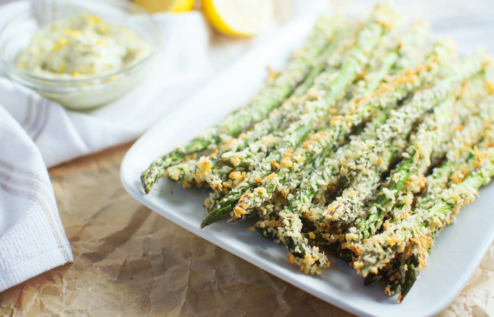A plate of crispy Asparagus Parmesan Fries coated in golden breadcrumbs sits on a white dish, with a bowl of creamy dip and lemon wedges in the background.