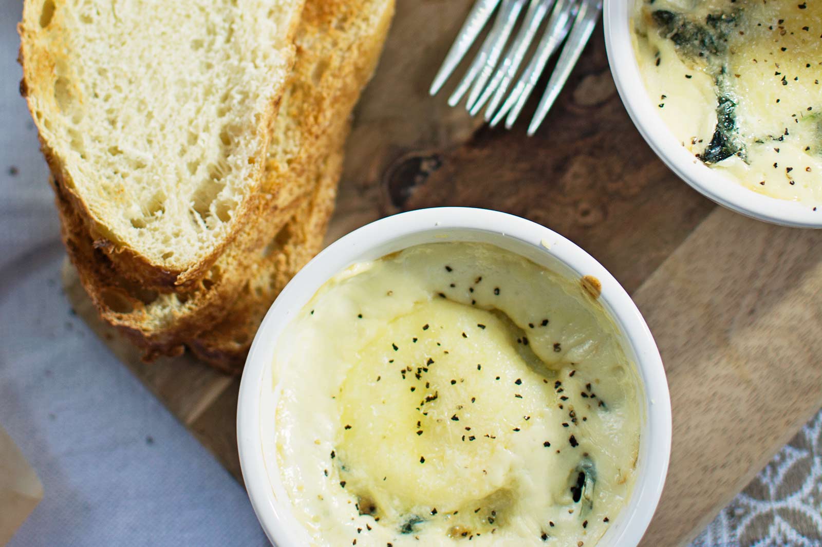 A close-up of a baked brunch dish in a white ramekin, topped with melted cheese and black pepper, next to slices of rustic bread on a wooden board, with a fork nearby.