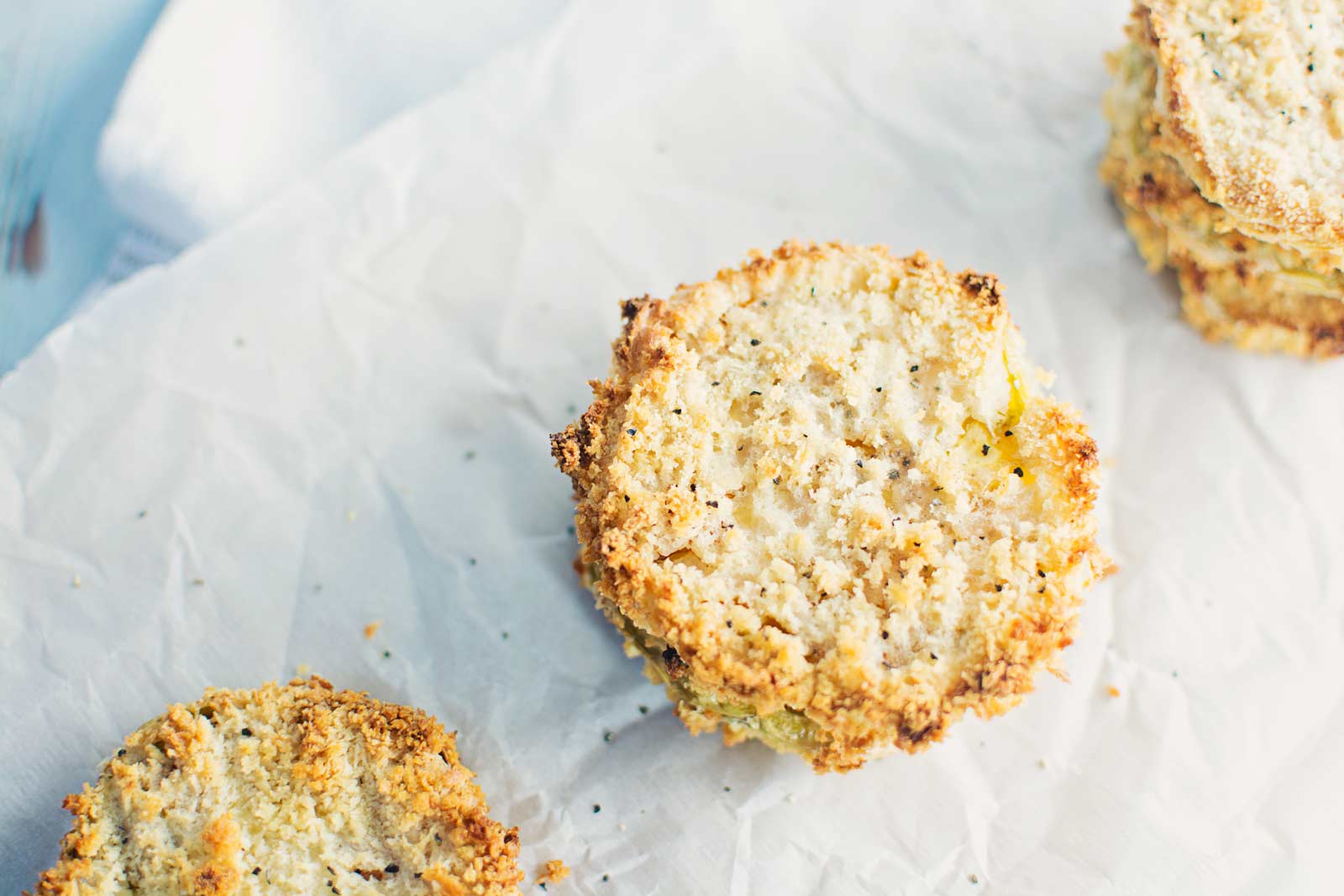 Three crispy, round, breaded and baked slices of food, possibly green tomatoes or eggplant, are placed on a sheet of white parchment paper, with a light and airy texture visible on their golden-brown edges.