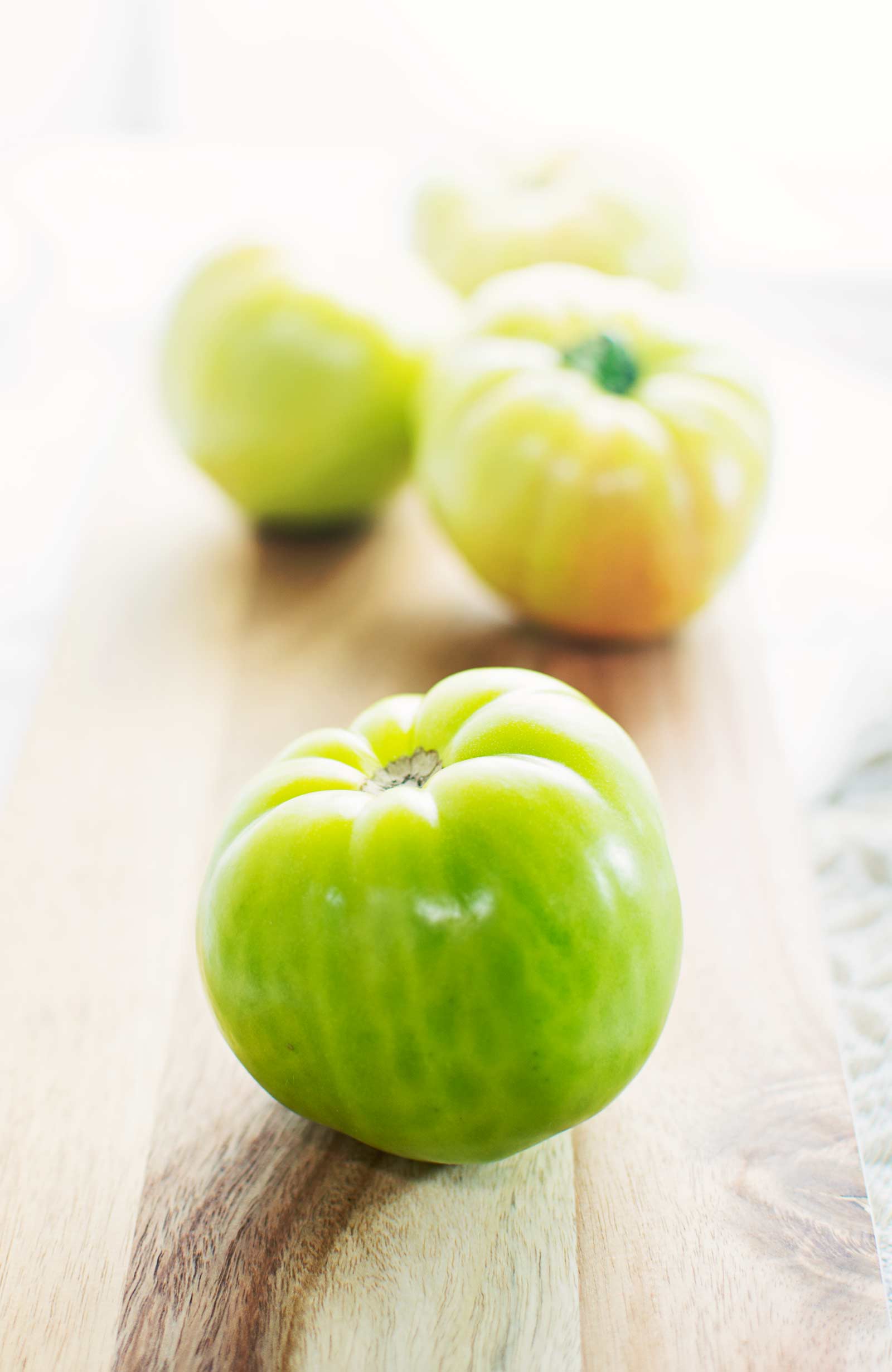 Four green tomatoes sit on a light wooden board, with one green tomato in sharp focus at the front and the others softly blurred behind. Bright, natural light highlights these fresh green tomatoes.