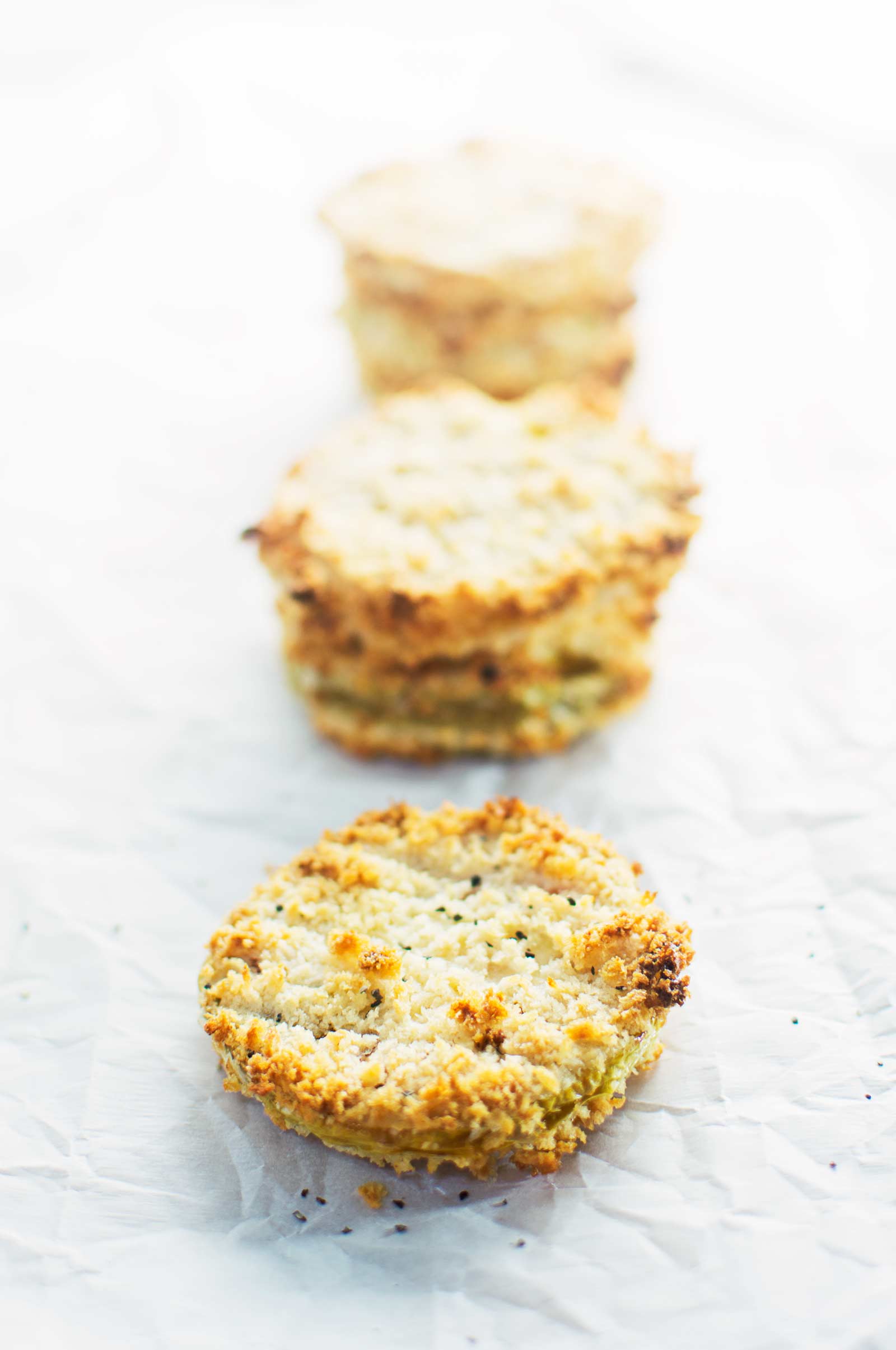 Four golden brown, round cauliflower patties are lined up on white parchment paper, alongside slices of fresh green tomatoes. The closest patty is in sharp focus while the others and the tomatoes fade gently into the background.