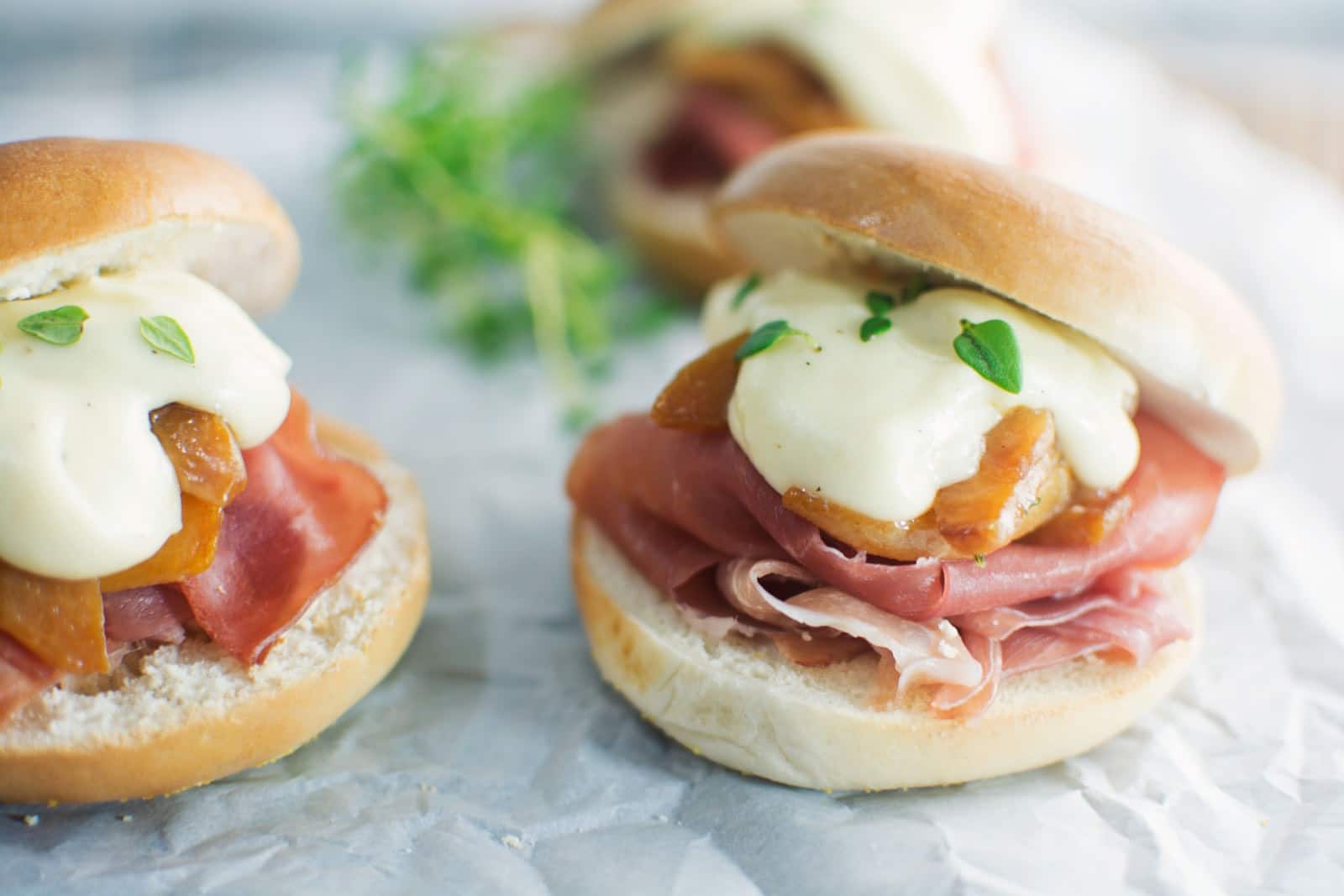 Three small bagel sliders with prosciutto, caramelized onions, and creamy white sauce on soft buns, garnished with fresh green herbs, sit on parchment paper.