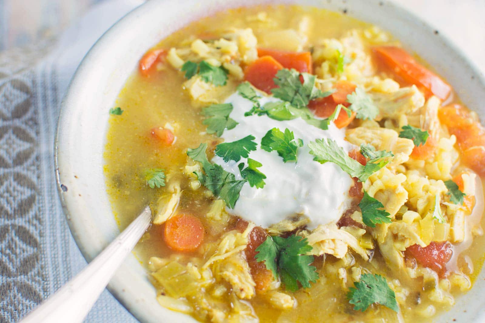 A comforting bowl of Chicken Soup with carrots, rice, and shredded chicken, topped with a dollop of yogurt and fresh cilantro. A spoon rests in the hearty soup, ready to enjoy.