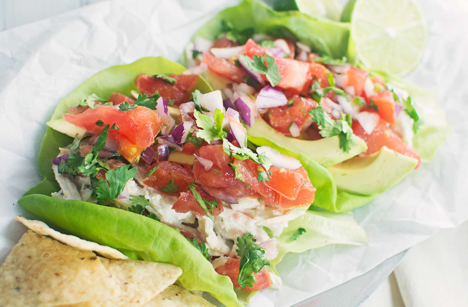 Leafy lettuce wraps filled with flavorful chicken salad, diced tomatoes, red onions, avocado, and cilantro, served with tortilla chips and lime wedges on a white surface.