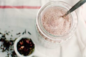 A glass jar filled with pink Hibiscus Sugar, with a spoon inside. Next to it is a small white dish containing dried herbs and flower petals, all placed on a white cloth with a red stripe.
