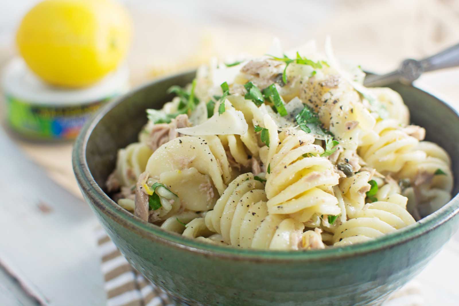 A green bowl filled with rotini pasta mixed with tuna, herbs, and shredded cheese, topped with black pepper. In the background, a can of tuna and a lemon are softly blurred.