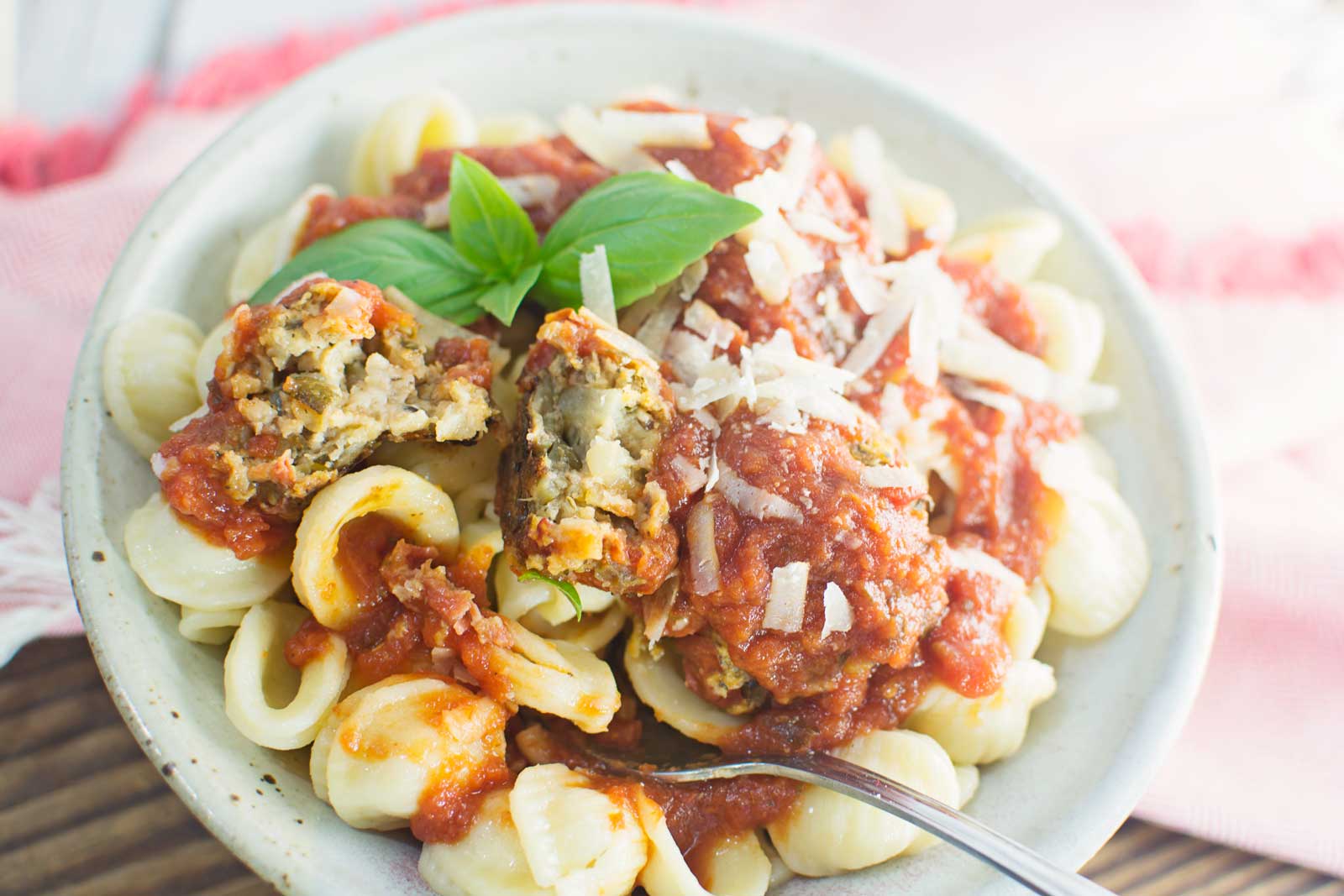 A bowl of shell pasta topped with tomato sauce, grated cheese, and Veggie Balls, garnished with fresh basil. A fork and a halved Veggie Ball are visible, revealing the delicious inside texture.
