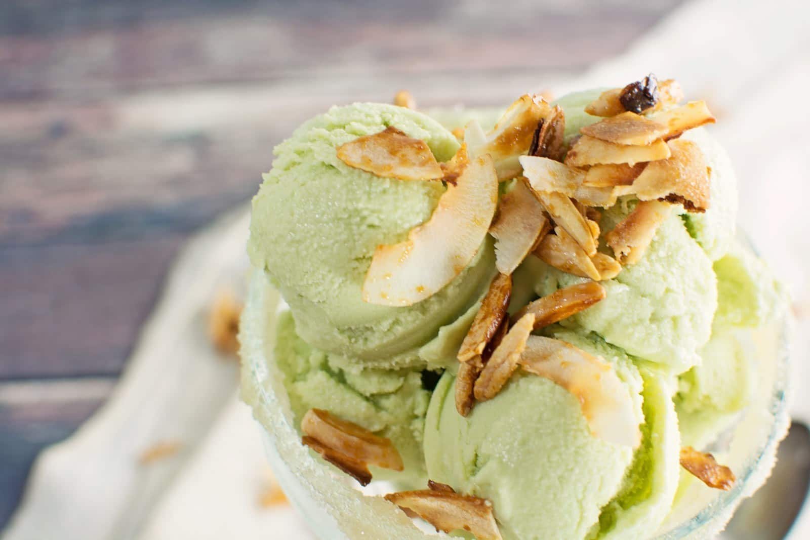 Scoops of green pistachio and avocado ice cream topped with toasted almond slices in a glass bowl, set on a light surface with a blurred background.