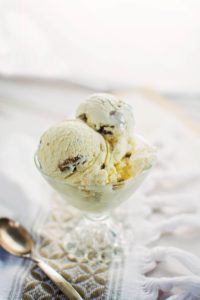 A glass dessert dish filled with two scoops of vanilla ice cream with chocolate chunks and a hint of butter pecan ice cream, placed on a textured cloth next to a silver spoon.