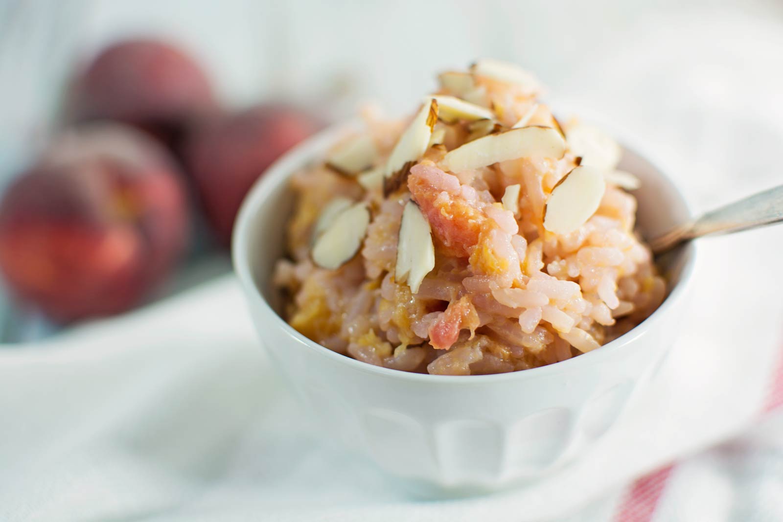 A white bowl filled with creamy rice pudding, topped with peach pieces and sliced almonds. A spoon rests in the bowl, while peaches are blurred in the background on a white surface—a sweet twist far from classic fried rice.