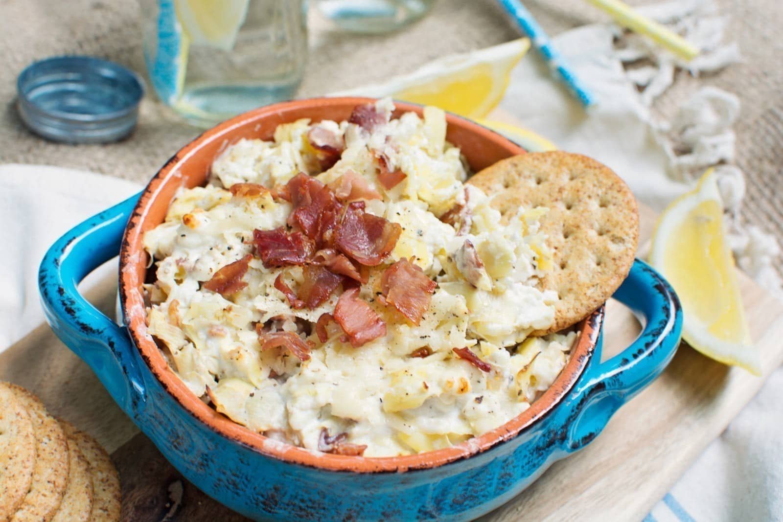 A blue bowl filled with creamy Prosciutto Artichoke Dip topped with crumbled bacon, served with a round cracker. Lemon wedges, crackers, and a glass of water are nearby on a wooden surface.