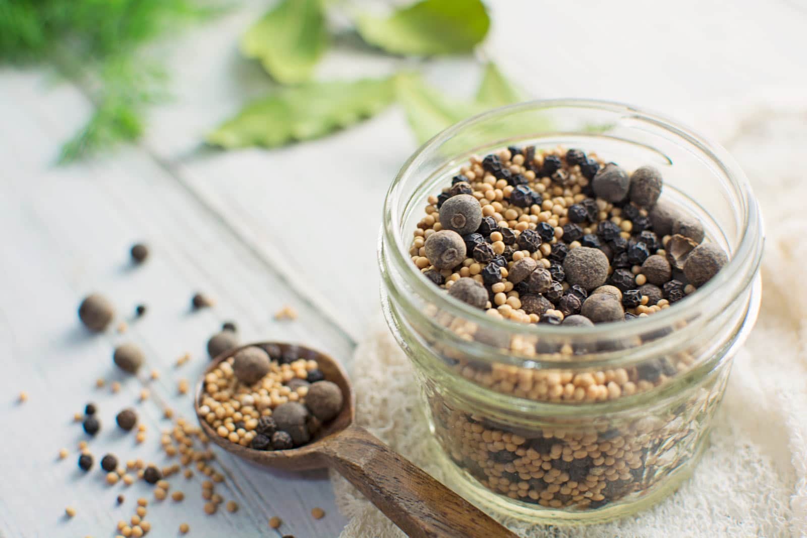 A glass jar filled with a mix of whole pickling spice, including mustard seeds and peppercorns, sits on a light surface. A wooden spoon with more spice mix and scattered seeds is beside the jar. Green herbs are in the background.