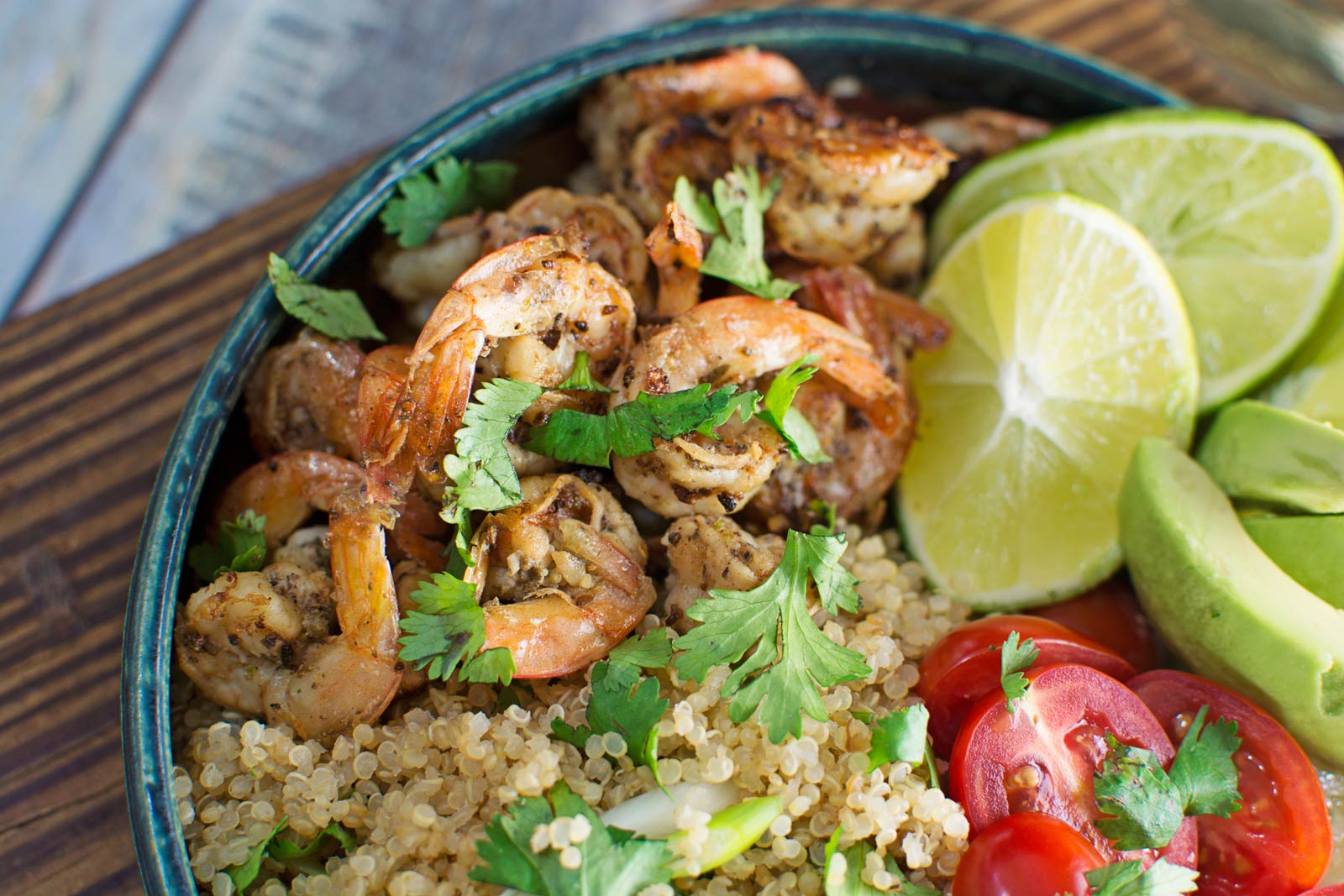 A bowl with Garlic Lime Shrimp, quinoa, sliced cherry tomatoes, avocado, lime wedges, and fresh cilantro on top, presented on a wooden surface.